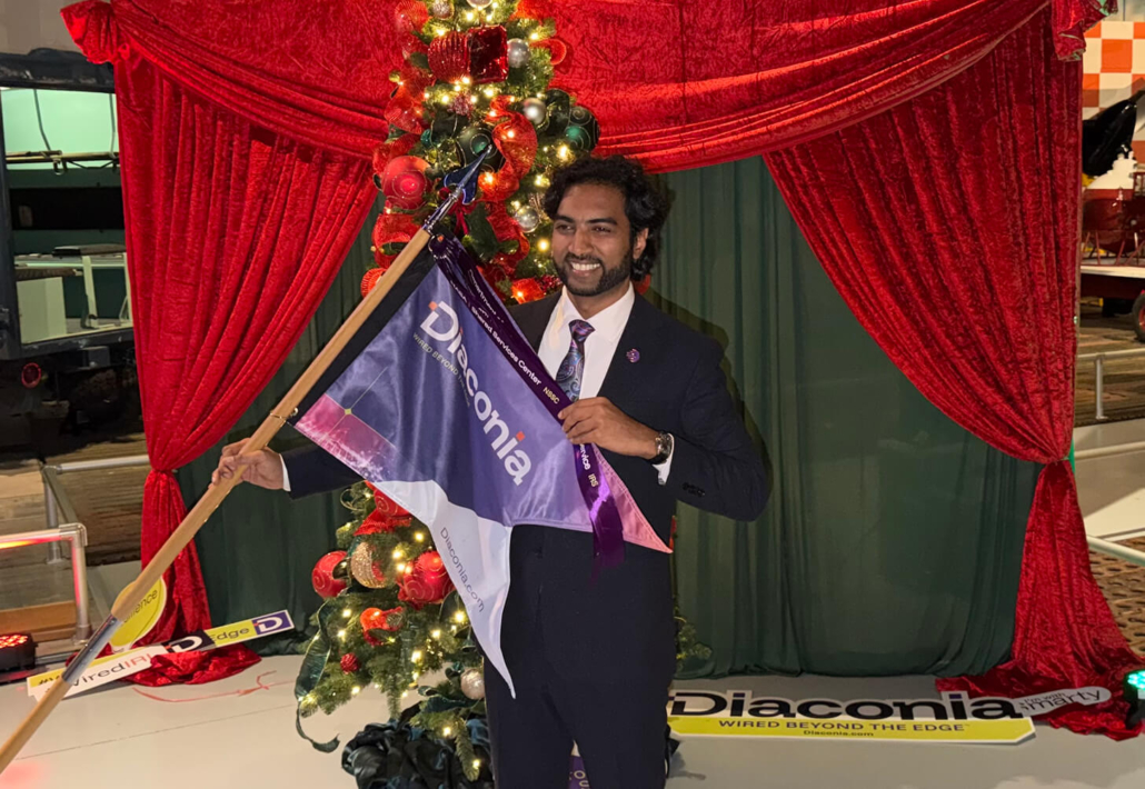 A man in a suit holding a Diocesan flag stands in front of a decorated Christmas tree with red curtains and festive lights.