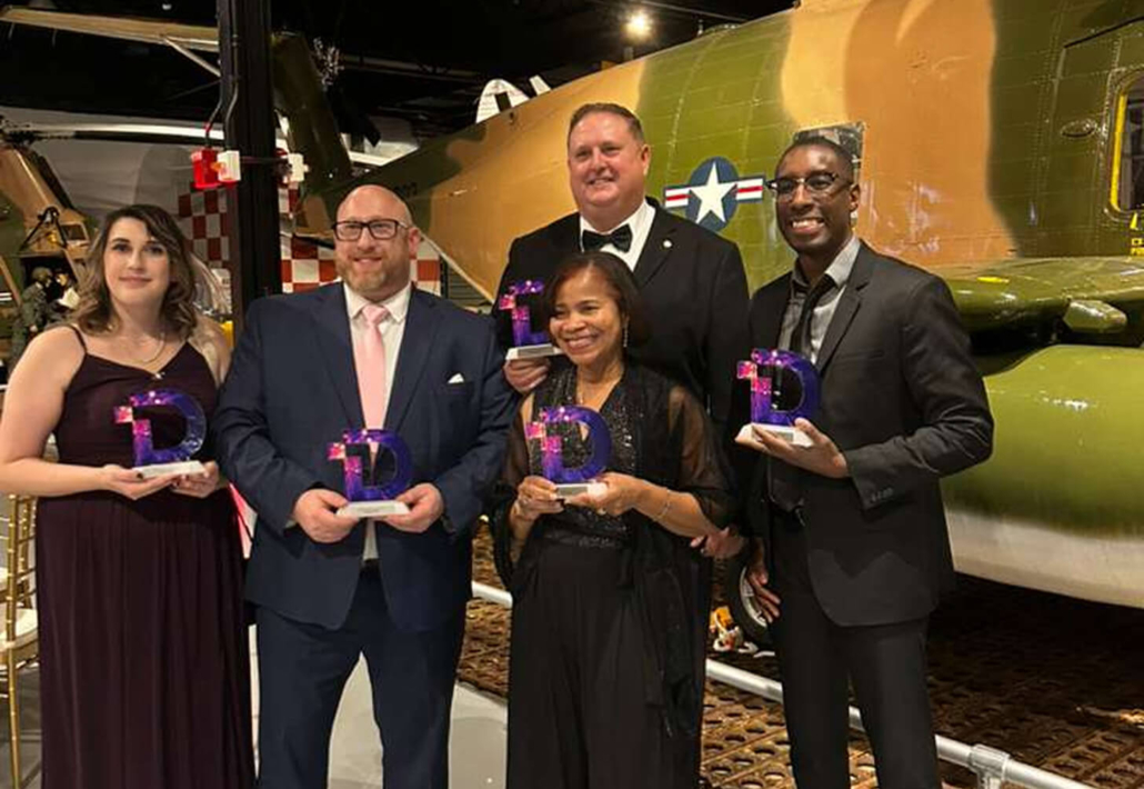 Five people in formal attire holding awards with a vintage aircraft in the background at a celebration event.