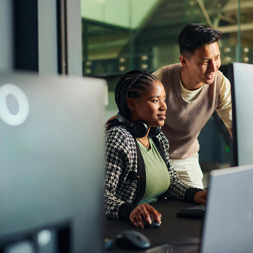 A woman with headphones and a man assist her at a computer in a modern office, illustrating AI and decision support tools.