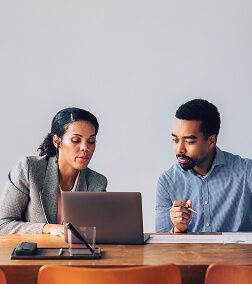 Two professionals work together at a desk with a laptop, discussing documents in a modern office setting.