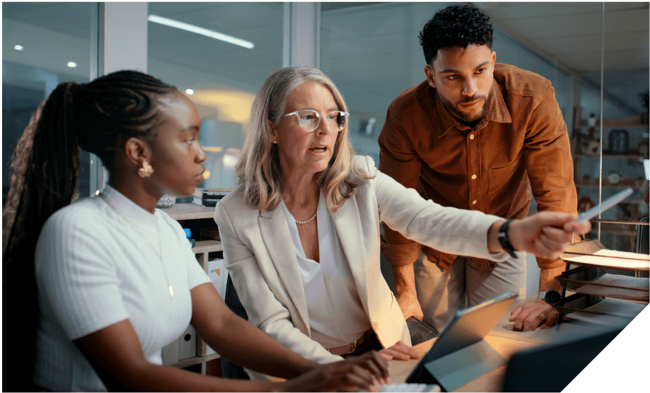 Three professionals collaborate at a desk, with one woman pointing at a laptop screen during a discussion.