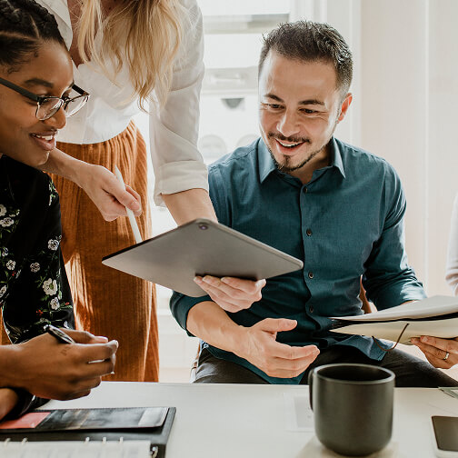 Three diverse professionals collaborate around a tablet and documents in a bright office space.