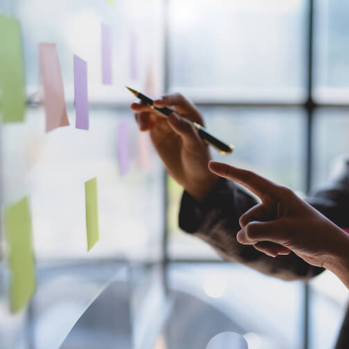 Hands point and write on colorful sticky notes on a glass wall during a brainstorming session.