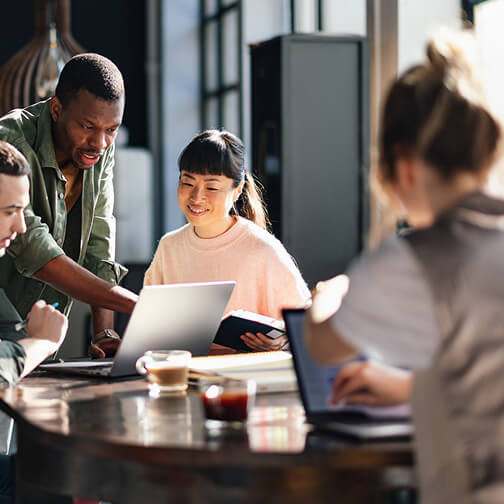 A diverse group of people collaborates around a table with laptops, books, and coffee in a bright, modern workspace.