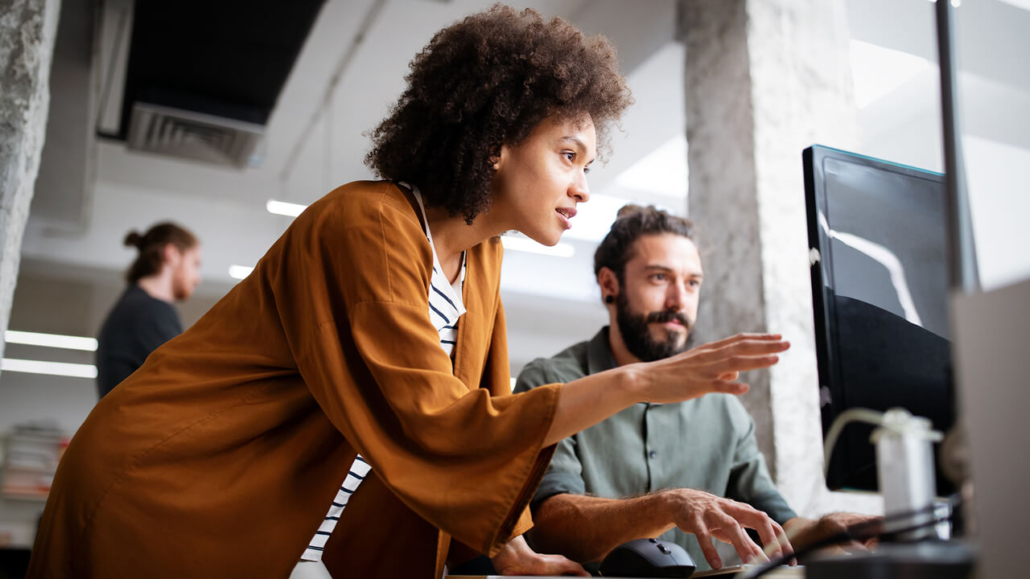 A woman in a brown jacket guides a man at a computer, both focused on the screen in a modern office setting.