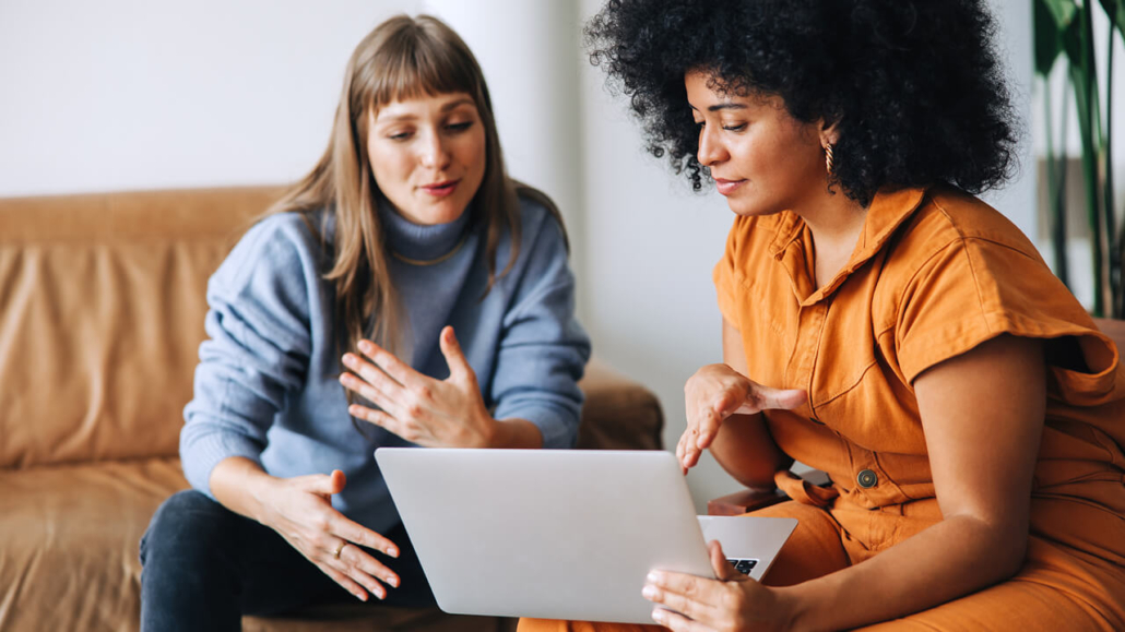 Two women discuss security concerns while looking at a laptop, with one gesturing towards her chest and the other pointing