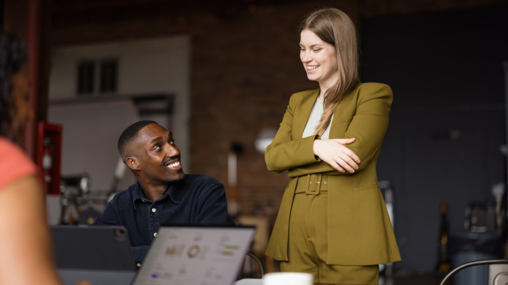 A woman in a green suit smiles and talks with a man sitting at a table, engaging in a friendly conversation in an office