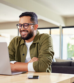 A smiling man with glasses and a beard sits at a desk with a laptop, in a bright modern office setting.