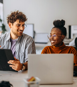 Two smiling young adults, one with curly hair and glasses and the other with a bun and glasses, work together at a laptop.