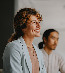 Smiling woman with curly hair in a light blazer, sitting next to a man with long hair tied back, in a professional setting.