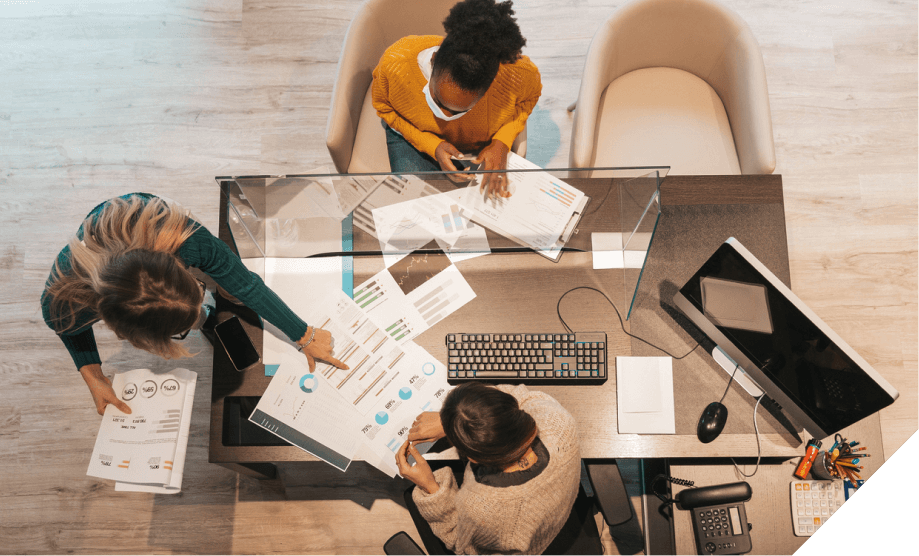 Three professionals collaborate at a desk with charts, a computer, and documents, focusing on data analysis and applied