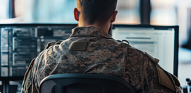 A person in military uniform works on multiple computer screens displaying code and data.