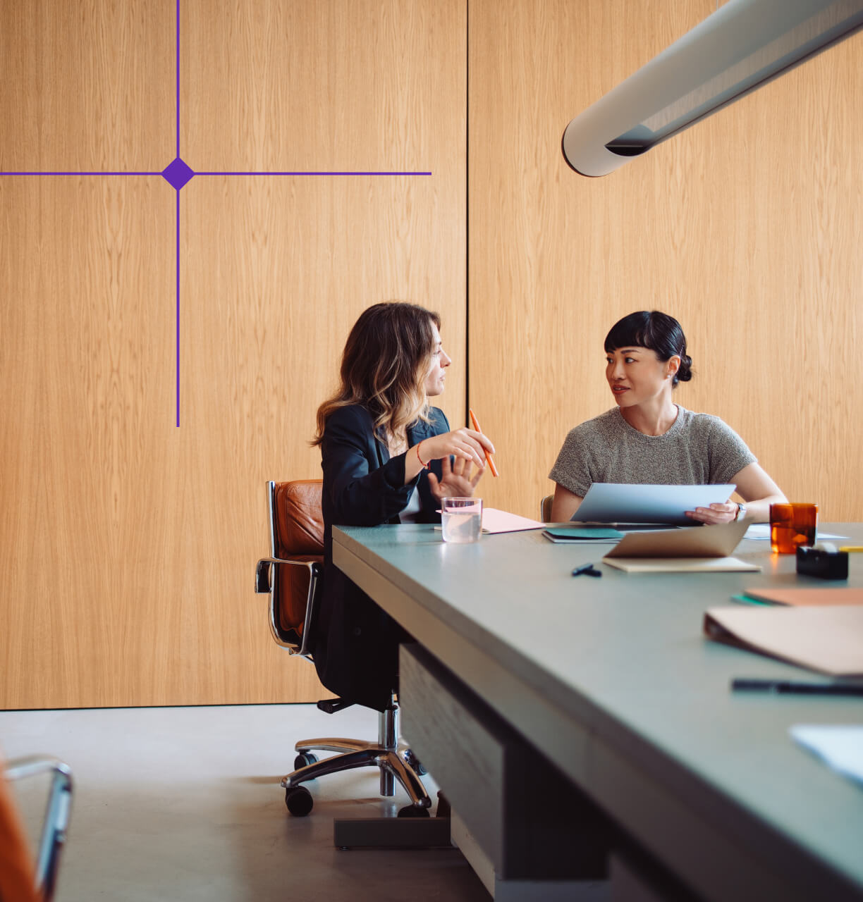 Two women sit at a table in a modern office, engaged in a conversation with documents and a glass of water on the table.