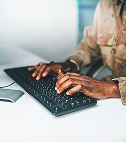 A person types on a keyboard with a notebook and glasses nearby on a desk.