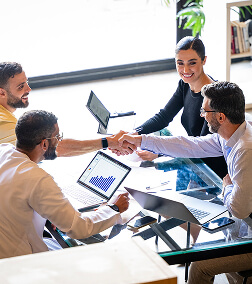 A woman in a black blazer shakes hands with a man across a table, surrounded by colleagues with laptops in a bright office.
