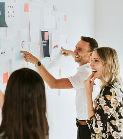 Three people smiling and pointing at a wall with papers and notes during a discussion or presentation.