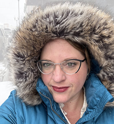 A woman with glasses wearing a blue jacket with a fur-lined hood outdoors in cold weather.
