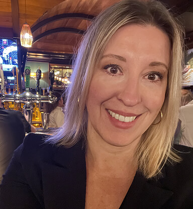 A smiling woman with blonde hair in a dark blazer, in a lively indoor setting with warm lighting and bar elements in the