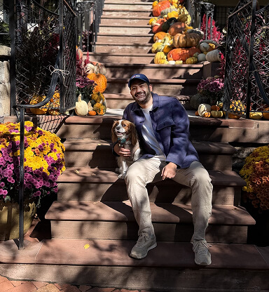 A man with a dog sitting on outdoor steps decorated with pumpkins and colorful flowers for fall.