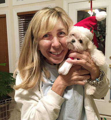 A smiling woman with blonde hair holding a small white dog wearing a Santa hat in a cozy indoor setting.