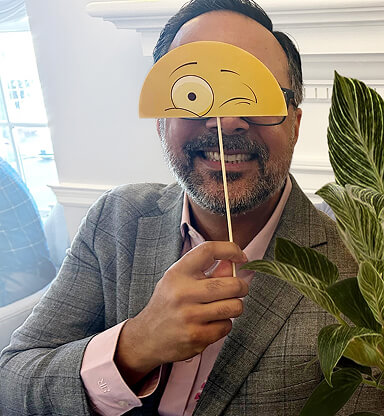 A man in a gray suit holds a yellow, winking emoji mask on a stick in front of his face, smiling behind it.