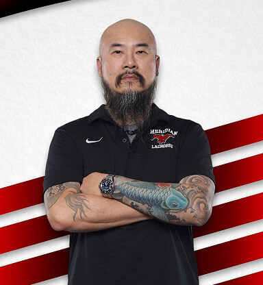 A confident man with a beard and tattoos on his arms, wearing a black polo with a sports logo, stands with arms crossed