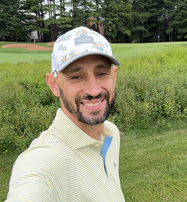A smiling man with a beard wearing a white cap with floral patterns and a light yellow striped shirt stands outdoors on a