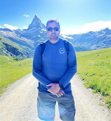A man wearing sunglasses and a blue jacket stands on a mountain trail with green hills and snow-capped peaks in the