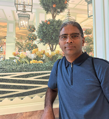 A man with glasses in a blue polo shirt stands indoors near a window with a decorated indoor garden and Christmas ornaments