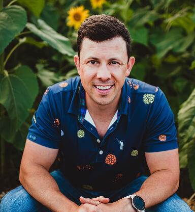 A smiling man with short dark hair wearing a blue patterned shirt and a smartwatch, sitting outdoors among green plants.