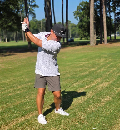 A man in a hat and sunglasses swings a golf club on a sunny golf course with trees in the background.