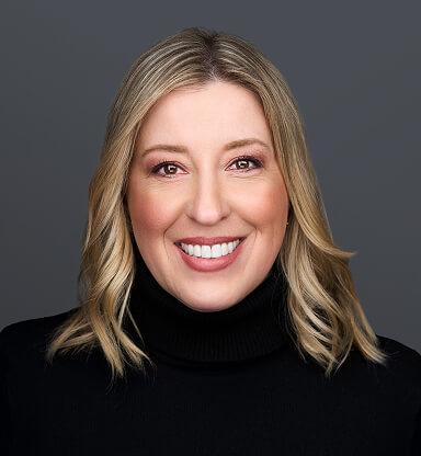 A smiling woman with blonde hair wearing a black turtleneck against a gray background.
