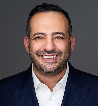 A smiling man with short dark hair, a beard, wearing a white shirt and dark blazer against a gray background.