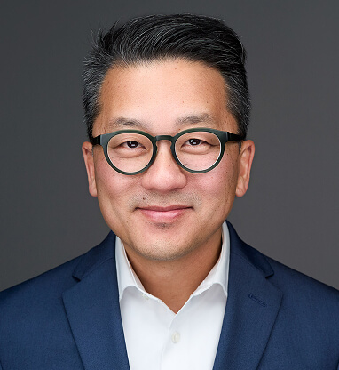 Jonathan Chu wearing glasses and a navy blazer, smiling against a gray background.