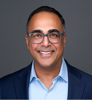 A smiling man with glasses, wearing a blue suit and light blue shirt, poses against a gray background.