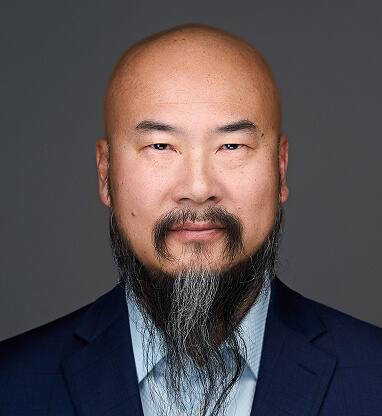 A professional headshot of Patrick Kuo with a bald head, beard, and mustache, wearing a dark suit and light blue shirt.