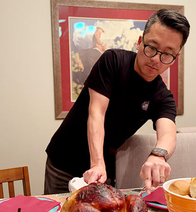 A man with glasses and a black shirt carves a roasted turkey at a dining table.