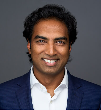 A smiling man with dark hair, wearing a navy blazer and white shirt, against a gray background.