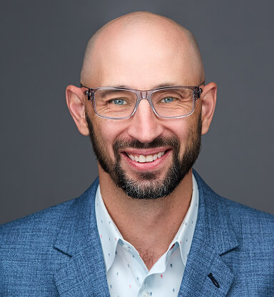 A smiling, bald man with glasses, wearing a blue blazer and white shirt, poses against a gray background.