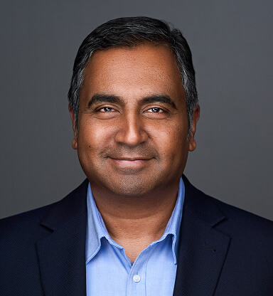 A professional headshot of Santosh Srinivasaiah in a dark blazer and light blue shirt, smiling against a gray background.
