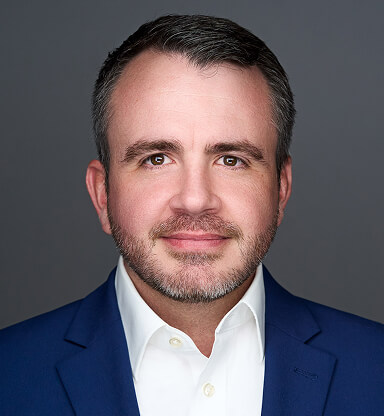 A professional headshot of Scott Bancroft in a blue suit and white shirt against a gray background.