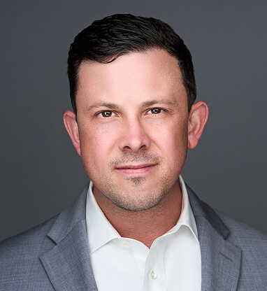 A professional headshot of Tom Adams in a gray suit and white shirt against a plain gray background.