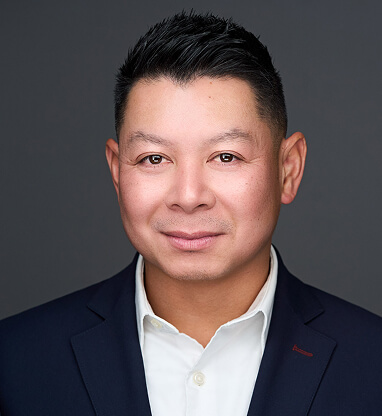 A professional headshot of Tom Simanivanh in a dark suit and white shirt against a gray background.