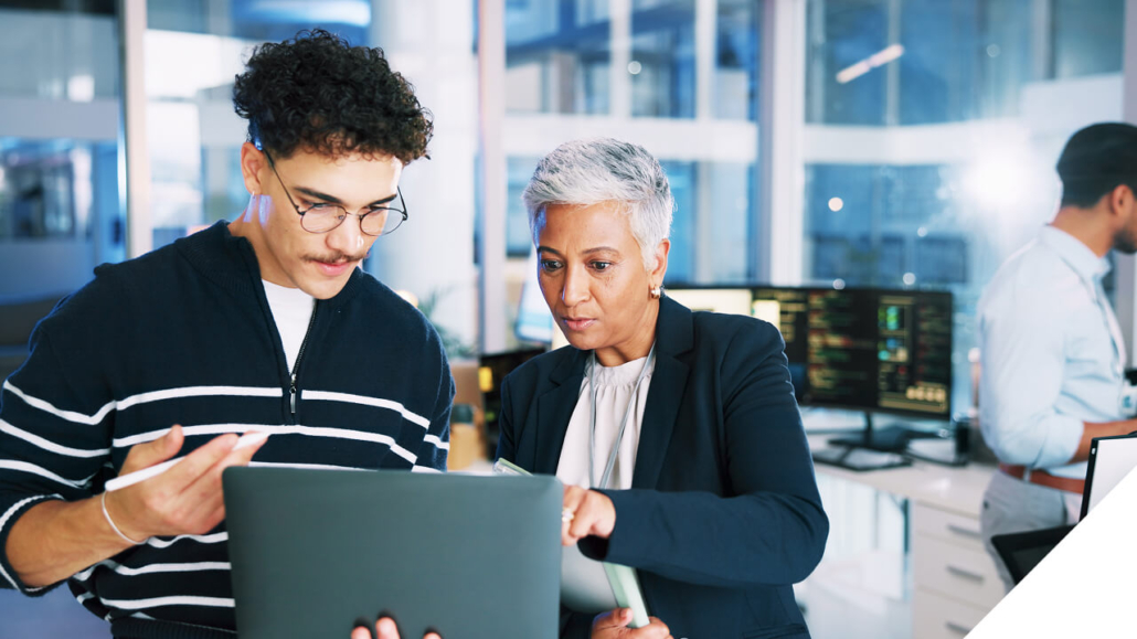 Two professionals review a laptop screen in a modern office with multiple monitors and large windows.