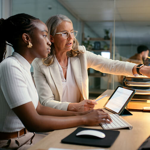 A woman and a young girl collaborate at a desk, looking at a tablet and computer screen in a professional office environment.