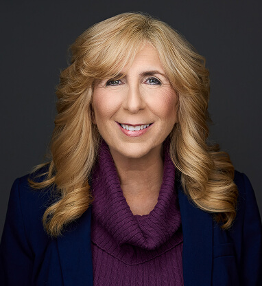A smiling woman with long blonde hair, wearing a purple turtleneck and a dark blazer against a gray background.