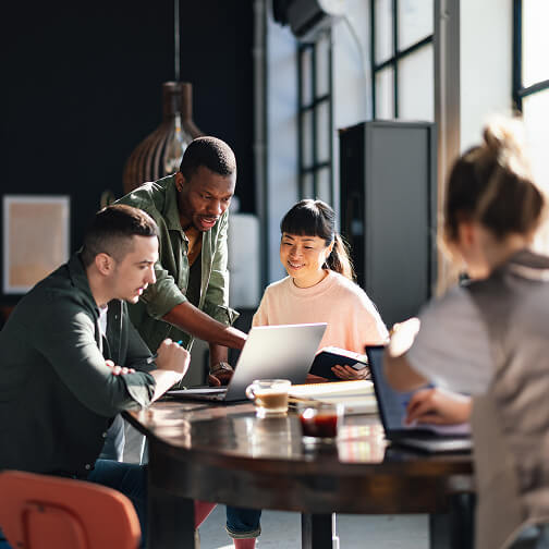 A diverse group of people collaborates around a table with laptops and tablets in a bright, modern office space.