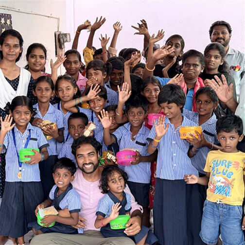 A group of smiling children and adults, some raising hands, gathered indoors with food containers, celebrating community and