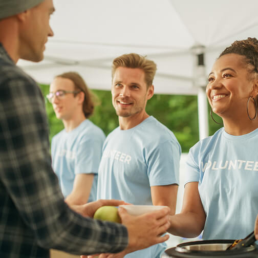 Volunteers in light blue shirts shake hands with a man at a community event under a tent.