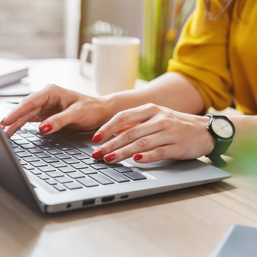 A woman typing on a laptop with a yellow shirt, watch, and red nail polish, sitting at a desk with a mug and books nearby.